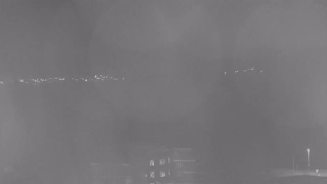 A monochrome image depicting a foggy or hazy view. In the distance, a faint line of lights is barely visible, suggesting a cityscape or roadway. The foreground is mostly dark, with vague outlines of structures and trees. Overall, the scene conveys a sense of low visibility and atmospheric conditions, creating a muted and unclear perspective.