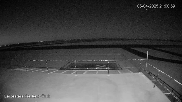 A dark, nighttime image captured from a webcam at Leicestershire Aero Club. The scene features a flat surface that appears to be a landing area, with some marked lines forming patterns on it. A railing runs along the foreground, slightly obscuring the view of the landing area. The background shows a faint horizon with distant lights illuminated, suggesting the presence of a city or populated area. The overall atmosphere is quiet and still, indicative of nighttime.