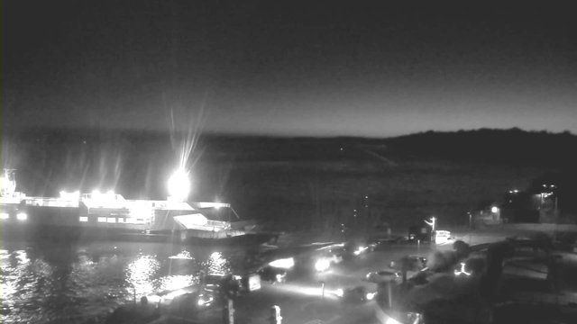 A dark, nighttime scene showing a harbor with a large illuminated boat docked on the left. The water reflects the lights of the boat and surrounding vehicles, while the shoreline features parked cars and faint outlines of nearby structures. The sky gradually lightens towards the horizon, suggesting dawn.