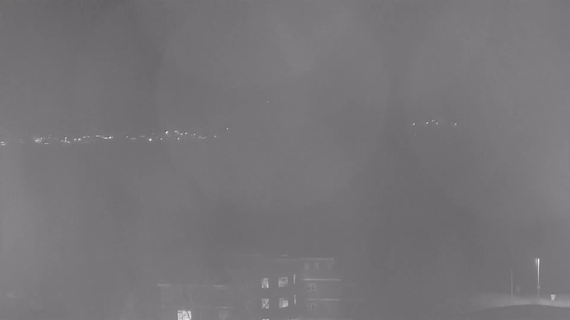 A foggy night scene with limited visibility. In the foreground, there is a dark building with several windows emitting a faint light. In the background, a faint line of lights is visible, suggesting a distant horizon or road, but details are obscured by fog. The overall appearance is muted and monochromatic, primarily in shades of gray.