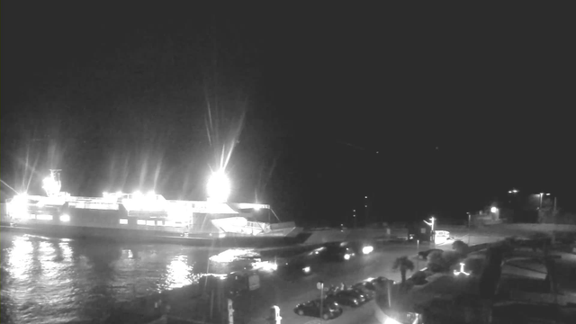 A brightly lit ferry is docked at a harbor at night, with its lights reflecting on the calm water. In the foreground, there are parked vehicles, and in the background, there are faint outlines of distant structures and lights.