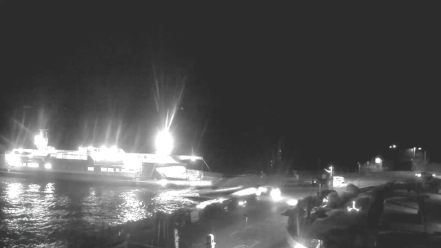 A nighttime scene featuring a brightly lit ferry docked at a pier. The ferry is illuminated, with strong lights shining down, reflecting off the water's surface. In the foreground, there are dark silhouettes of vehicles and structures, with a faint outline of a road leading away from the dock. The background is mostly dark, suggesting an evening atmosphere with minimal visibility.