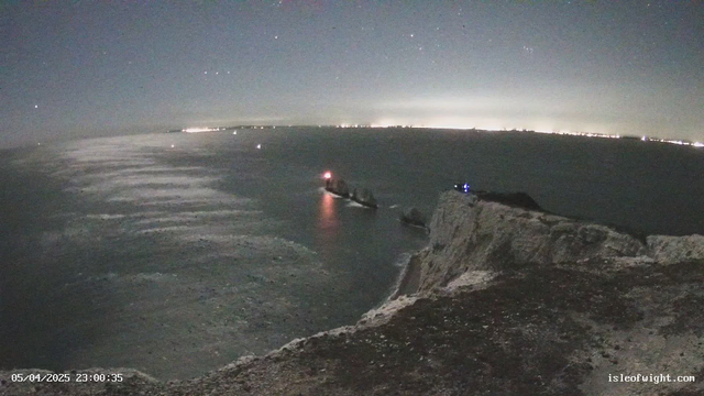A nighttime scene of the ocean viewed from a cliff. The water reflects a shimmering light, indicating the presence of waves. In the foreground, the rugged cliff edge is visible, leading into the dark water. To the left, there are two boats, one displaying a red light, possibly a navigation or warning light. The background shows distant city lights along the horizon, creating a glow against the dark sky, which is filled with stars. The date and time stamp is located at the bottom left corner of the image.