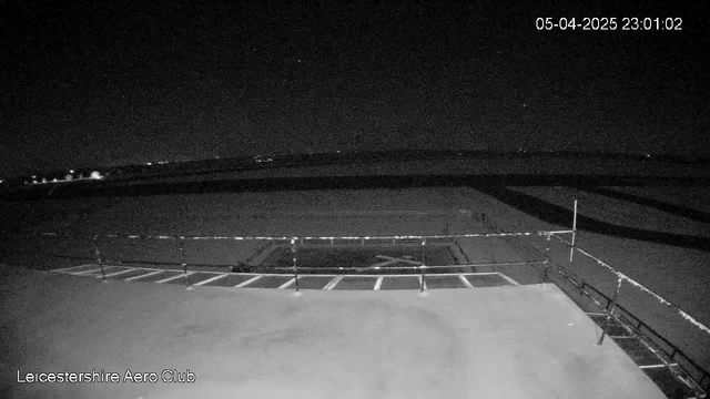 A dimly lit scene showing a flat rooftop with railings. The foreground features a section of a flat surface, possibly concrete, with a ladder-like structure and white lines suggesting positions or markings. In the background, faint lights from distant structures or vehicles are visible against a dark sky, indicating a nighttime setting. The date and time at the top right corner display "05-04-2025 23:01:02." The overall ambiance is quiet and still, with limited visibility.