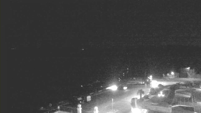 A dark, nighttime scene captured by a webcam. The foreground shows a road with several vehicles, and a few light sources illuminating the surroundings. On the left, there are structures, possibly buildings or streetlight poles, and a subtle glow in the background indicating some distance lights, suggesting the presence of water or horizon. The image is predominantly dark with minimal visibility.