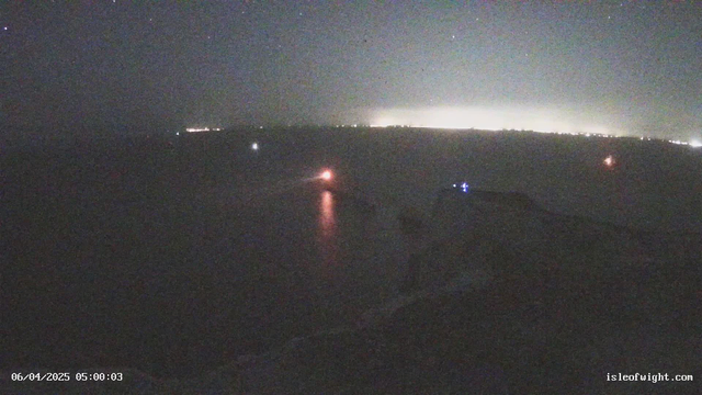 A dimly lit coastal scene at night, featuring a body of water reflecting faint light. A small boat emits a red light, while another, further in the distance, glows blue. Soft white lights from the shoreline suggest buildings or structures. The sky is dark with scattered stars, and there is a haze near the horizon, indicating city lights beyond the water. The date and time displayed at the bottom left are June 4, 2025, at 05:00:03.