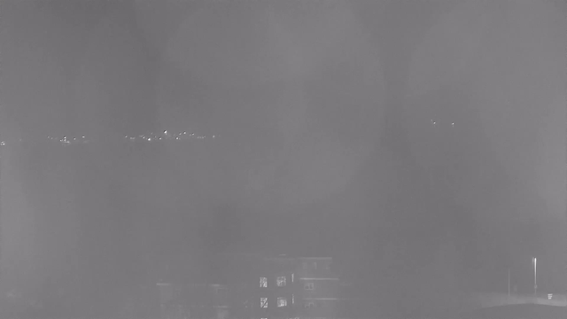 A dimly lit webcam image captures a hazy scene. In the foreground, there are dark silhouettes of buildings with a few faint illuminated windows. In the background, a blurred line of lights is visible, suggesting a distant horizon or streetlights. The overall tone is gray, indicating low visibility, likely due to fog or weather conditions.