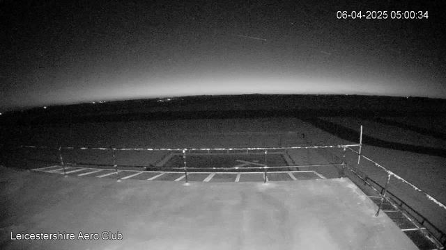 A dark scene captured from a webcam showing an expansive view of the sky and horizon. The ground appears flat with white lines marking where vehicles would park. A low railing is visible in the foreground, and there are faint lights in the distance. The background gradually transitions into a lighter grey at the horizon, suggesting dawn or dusk. The date and time are displayed in the upper right corner.