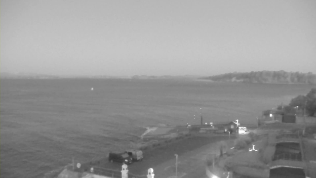 A black and white webcam image shows a calm body of water, stretching out towards distant landmasses that are barely visible. In the foreground, there is a small harbor area featuring a winding pathway, a parked vehicle, and light fixtures. The shoreline is lined with structures, including a building on the right side with lights illuminated. The overall atmosphere is serene and quiet, with gentle waves visible on the water's surface.