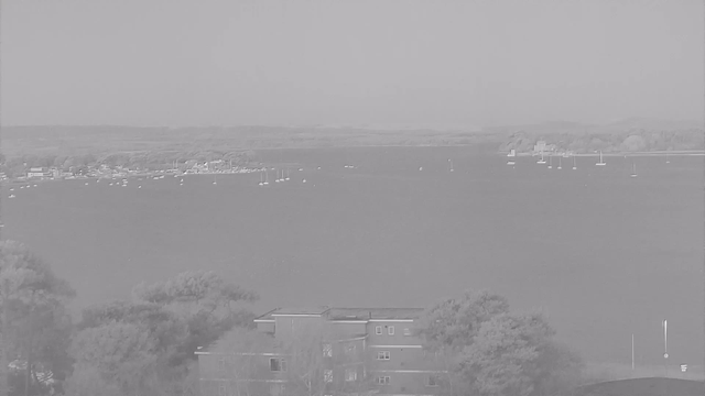 A gray-scale image showing a calm body of water with several small boats scattered across the surface. In the foreground, there are trees and a low building with multiple windows. The background features a distant shoreline with trees and possibly more structures, all leading towards a cloudy sky. The scene conveys a serene, tranquil atmosphere.