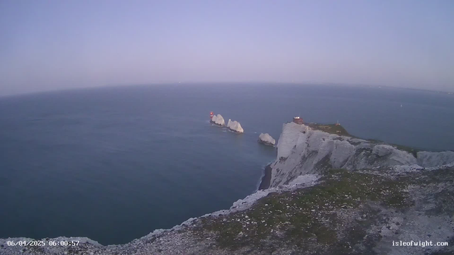 A view of a coastal landscape at dawn, featuring rugged white chalk cliffs on the right side of the image. In the water, there are several gray rock formations, with a red lighthouse positioned on one of the rocks. The sky is light blue, transitioning to pale pink near the horizon, and the calm sea reflects the colors of the dawn. A small building with a red roof is located at the top of the cliff, surrounded by green grass. The image captures a peaceful and serene atmosphere.
