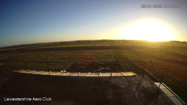 A view from a webcam overlooking the Leicestershire Aero Club. The scene depicts a wide, open field under a clear sky at sunrise, with the sun rising in the background. The ground is green with patches of grass and some visible markings. In the foreground, there is a low railing and concrete platform. The time displayed at the top shows 07:00:05 on April 6, 2025.