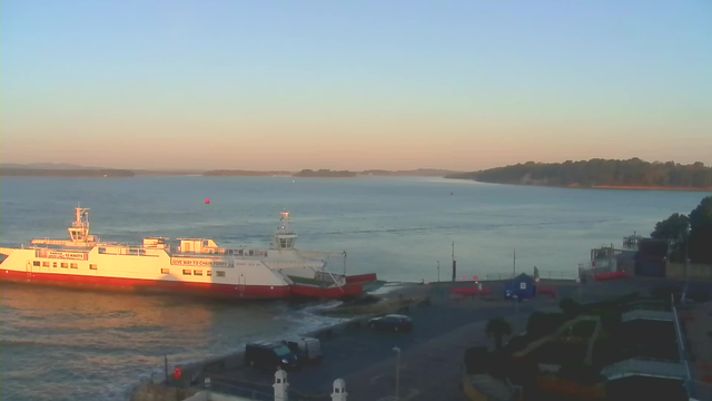 A ferry boat docked near a shoreline at sunset. The ferry is white with red accents, displaying a sign that reads "GIVE WAY TO CHAIN FERRY." In the background, the water is calm, reflecting soft colors of the sky—pale blues and warm oranges. Several small boats can be seen in the distance, along with green hills lining the opposite shore. The area in front of the ferry includes parked vehicles and landscaping, with a few palm trees visible.