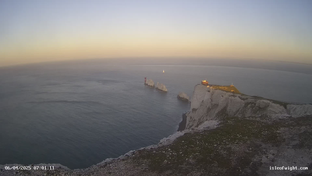 A coastal scene at sunrise featuring white cliffs on the right side and a calm sea reflecting the soft light. In the distance, a small sailboat is moving on the water. Two prominent rock formations, one with a red lighthouse, jut out into the sea. The sky displays a gradient from light blue to soft orange, indicating dawn. The foreground is grassy with visible rocky textures.