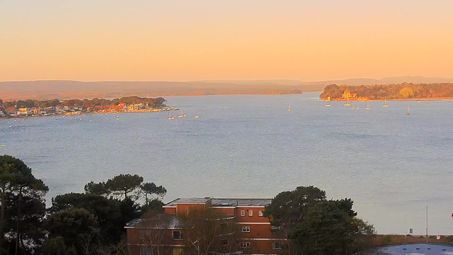 A serene waterfront view at sunset, featuring a body of water with multiple small sailboats scattered across. In the foreground, green trees and a brick building are visible, while across the water, a small town with buildings and a coastline can be seen. The sky exhibits warm hues of orange and yellow, creating a tranquil atmosphere. Gentle hills are present in the background, enhancing the peaceful scenery.