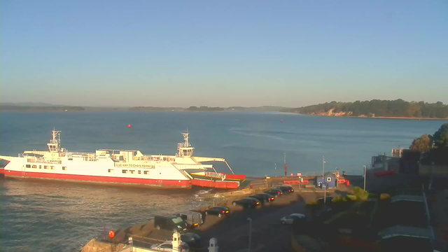 A ferry is docked at a terminal, with a calm body of water in the background. The sky is clear and blue, and there are small hills visible in the distance. Several cars are parked along the shoreline, and a few structures can be seen near the dock. A red buoy floats in the water. The overall scene conveys a tranquil morning by the waterfront.