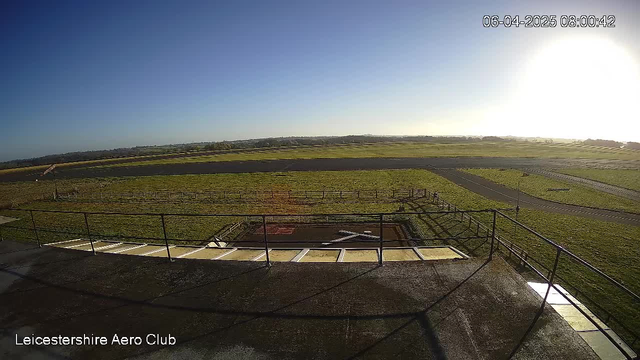A view from a webcam at Leicestershire Aero Club, showing a clear blue sky with the sun shining in the top right corner. Below, there is a green grassy area and a runway visible in the distance. A small structure appears near the center, and a few trees can be seen along the horizon. The image has a railing in the foreground and a slight incline leading down to the runway.