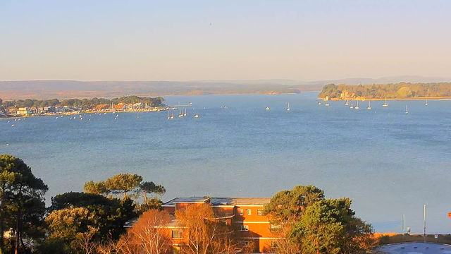 A scenic view of a calm blue water body surrounded by trees and buildings. In the foreground, there are several trees, while a building with a reddish exterior can be seen. In the distance, a shoreline features small boats scattered on the water, and a collection of houses with colorful rooftops line the shore. The backdrop consists of rolling hills under a clear sky. The atmosphere appears tranquil, with a warm light illuminating the landscape.