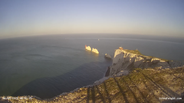A panoramic view of a coastline featuring white chalk cliffs under a clear blue sky. The sea is calm with gentle waves, reflecting the light. In the distance, there are two prominent rock formations, one with a red and white lighthouse structure, and the other smaller and more jagged. To the right, there is a building atop the cliffs, suggesting a lookout point. The foreground includes shadows cast on the rocky ground, indicating the time is early morning.