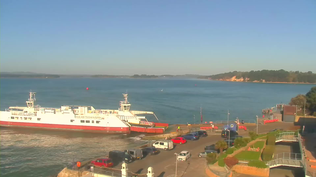 A large white ferry is docked at a waterfront, with its bow facing forward. The calm blue water extends towards the horizon, where green hills can be seen in the distance. In the foreground, a parking area is filled with various vehicles, including red and gray cars. Surrounding the parking lot are neatly maintained gardens and a small walkway that leads to a pier. The sky above is clear and sunny.