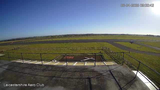 A view of an open field and runway from a webcam positioned at the Leicestershire Aero Club. The sky is clear and bright blue, with the sun shining in the upper right corner. In the foreground, there is a balcony with a railing, leading to a grassy area below. The runway stretches across the middle of the image, surrounded by green grass and fenced areas. A white vehicle is visible on the gray runway, and there is a red and white windsock indicating wind direction. The scene conveys a sunny morning atmosphere.