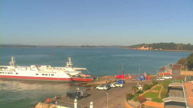 A calm waterfront scene featuring a large white and red ferry docked at a harbor. Several cars are parked nearby on a concrete area, with people walking around. The water is blue, reflecting the clear sky above, which has a few distant hills on the horizon. In the background, a sandy coastline with trees is visible. The overall atmosphere is peaceful and sunny.