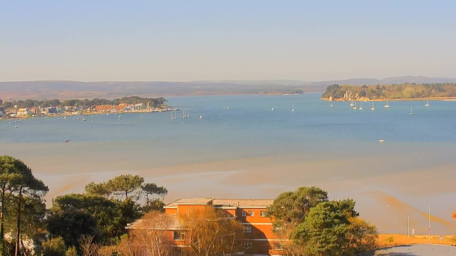 A serene view of a coastal landscape featuring a calm bay with several sailboats scattered across the water. In the foreground, there are some trees and a low building with an orange exterior. The background showcases a shoreline lined with houses and greenery, while hills are visible in the distance under a clear blue sky. The overall scene conveys a tranquil and picturesque seaside atmosphere.
