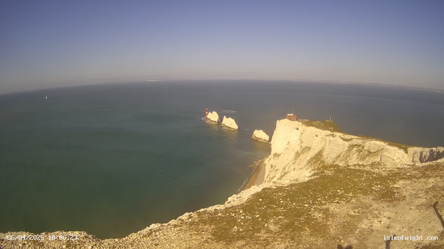 A coastal view featuring white chalk cliffs that drop into a calm blue sea. In the distance, there are two red and white lighthouses situated on rocky outcroppings. A small sailboat is visible in the water to the left of the lighthouses. The sky is clear and bright, transitioning from light blue to darker shades near the horizon. The foreground shows grassy terrain along the cliff edge.