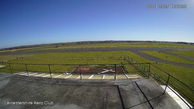 A clear blue sky stretches above a green landscape. In the foreground, a flat rooftop with a railing overlooks a grassy area and a runway. A helipad with white markings is visible on the left, and the grass is neatly maintained. To the right, a paved runway extends into the distance, with a few small roadways intersecting it. The scene conveys a peaceful, sunny day at an airfield.
