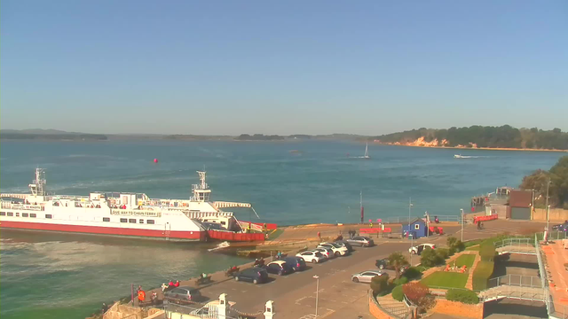 A serene view of a harbor under a clear blue sky. A large white ferry with red accents is docked at the left side, and several small boats can be seen on the water. On the right, there is a parking area with several cars, and a neatly landscaped area with greenery. In the distance, landforms hint at a coastal landscape with trees and cliffs. The scene conveys a peaceful maritime atmosphere.
