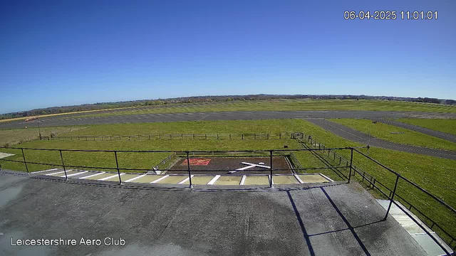 A clear blue sky with no clouds is seen above a grassy airfield. In the foreground, there is a flat rooftop area with a railing, and below it, a helipad marked with a red square and a white 'X'. The airfield stretches out with a few winding pathways and a fence bordering the grass. The date and time displayed in the top right corner indicate it is April 6, 2025, at 11:01 AM.