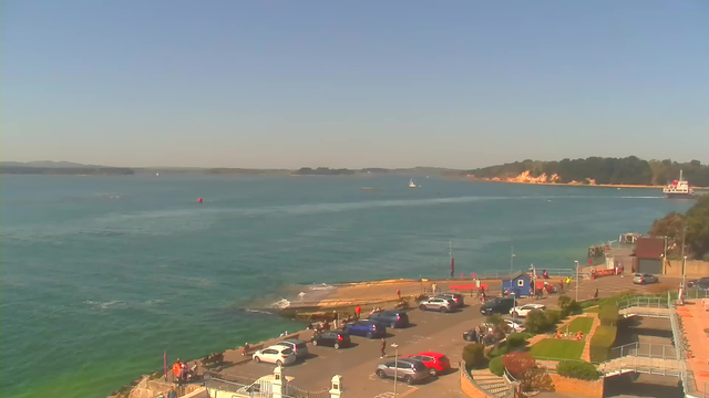A view of a seaside location on a clear day. In the foreground, there is a parking area filled with several cars. Some people are walking and gathering near the water's edge. To the right, a small harbor with a blue building and boats is visible. In the background, a sandy beach stretches along the coast with trees and a distant shoreline. The sky is mostly clear with a few wispy clouds, and the water is calm, featuring a gradient of blue and green tones.