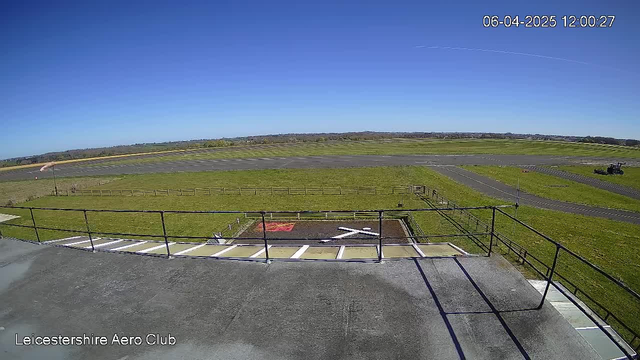 A clear blue sky is visible above an open grassy field, with a well-maintained runway stretching into the distance. In the foreground, there is a railing, indicating an elevated viewpoint, and a helipad marked with a white cross on a brown patch of earth. To the right, a tractor can be seen working on the grass. The landscape features a few trees and distant hills in the background. The timestamp on the image indicates the date and time.