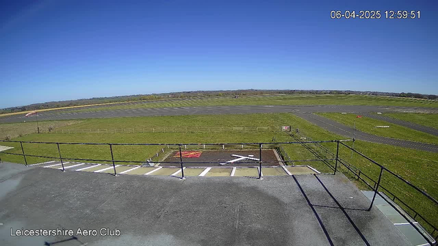 A clear blue sky is visible above an expansive green field and runway at an airfield. In the foreground, there is a railing along a platform, and markings indicating a runway position are visible on the ground, including a large white "X" among other markings. The scene is bright and sunny, with no visible aircraft in the area.