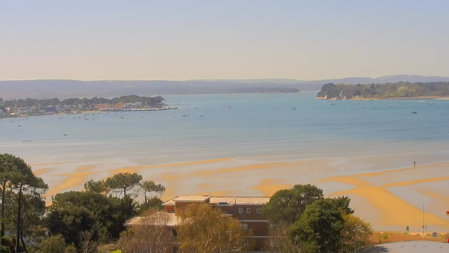 A serene coastal scene featuring calm blue waters and a sandy beach. Small boats are scattered across the water, with some moored near the shoreline. Lush green trees and shrubs are visible in the foreground, while a few buildings are positioned close to the beach. The distant land includes rolling hills under a clear sky. The sandy beach has areas where the tide has receded, revealing patterns in the sand.