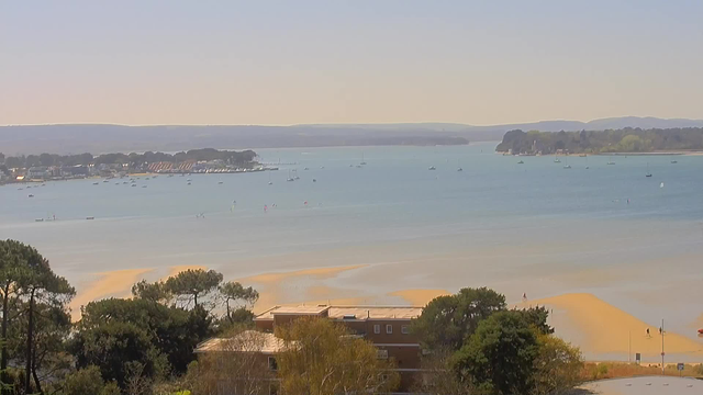 A serene coastal view captured from a webcam, featuring calm blue waters with several small boats floating. In the foreground, a sandy beach area is visible, framed by greenery and some buildings. The horizon is dotted with trees and distant hills, while the sky appears clear and bright, indicating a sunny day.
