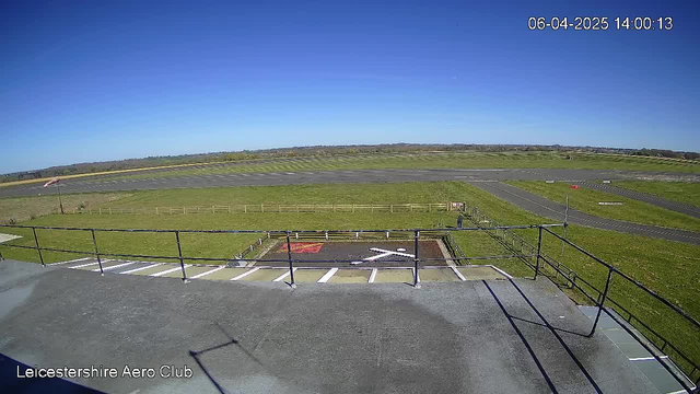 A clear blue sky stretches across the image, with bright sunlight illuminating the landscape below. In the foreground, a flat rooftop with a railing is visible, leading to a large open area featuring a grassy runway and outlines of grassy patches. On the runway, a small figure can be seen moving, and there are white markings on the ground, including a large cross shape. The background displays distant fields and hills, with a long stretch of asphalt running parallel to the grassy areas. A flag is positioned on the left, and traces of aircraft activity are apparent on the runway.