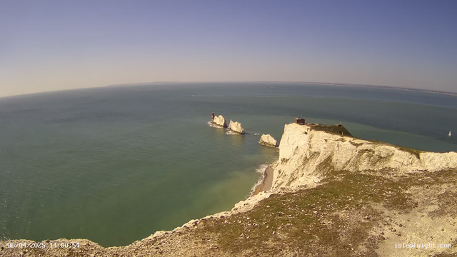 A panoramic view of a coastal landscape featuring white chalk cliffs on the right side, leading down to a sandy beach. Three rocky outcrops are visible in the water, with two small red structures atop them. The calm sea is a mix of blue and green hues, stretching out to the horizon under a clear sky. A small sailboat can be seen in the distance on the left side of the image. The image captures a tranquil, sunny day by the shore.