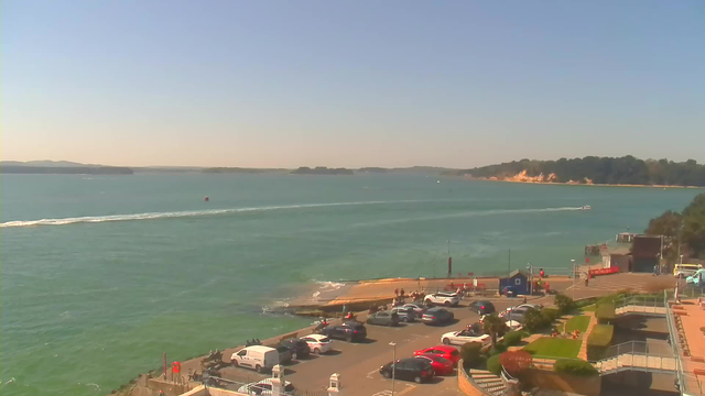 A coastal scene captures a clear blue sky and calm turquoise water. In the foreground, a parking lot filled with various cars is visible, alongside a concrete pier that juts into the water. A few people are seen walking near the water’s edge. In the background, a gentle slope leads to a wooded area and a sandy shoreline. A small boat is moving across the water, creating a wake. Farther out, distant landforms can be seen along the horizon.