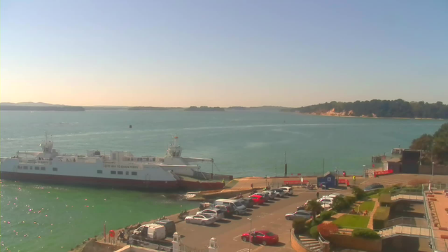 A sunny waterfront scene featuring a large ferry docked at a pier. The ferry is white with red detailing and has people on deck. In the foreground, there is a parking lot filled with cars of various colors. To the right, green grass and landscaped areas are visible, and further back, there are trees and a shoreline with sandy cliffs. The sky is clear and blue, reflecting sunlight on the calm water.