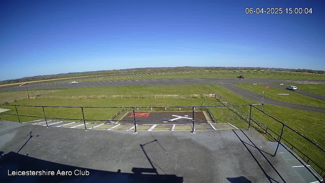 A clear blue sky is visible above a wide runway at the Leicestershire Aero Club. In the foreground, there is a fenced area with a marked helicopter landing zone featuring a white 'X' on the ground. A small airplane is seen taking off from the runway, and a vehicle is traveling along a path on the right side of the image. The landscape is green with a gently sloping background, and the overall scene conveys a sunny day at an airfield.