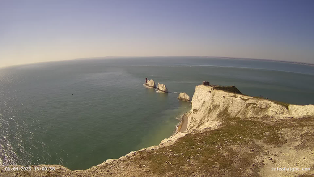 Wide view of a coastline featuring white chalk cliffs descending into a blue and green ocean. In the distance, two rock formations are visible in the water, with a small structure on one of them. The sky above is clear with a few clouds, and the sun is shining, creating a glimmer on the water's surface. There is a grassy area in the foreground, with people standing on the cliff edge. A sandy beach can be seen at the base of the cliffs.