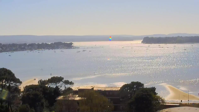 A serene waterfront view featuring a shimmering body of water under a clear blue sky. Small boats are scattered across the water, and a sandy shoreline is visible with a few people walking. In the distance, there are trees lining the shore and gentle hills or mountains beyond. The sunlight reflects off the water, creating a sparkling effect.
