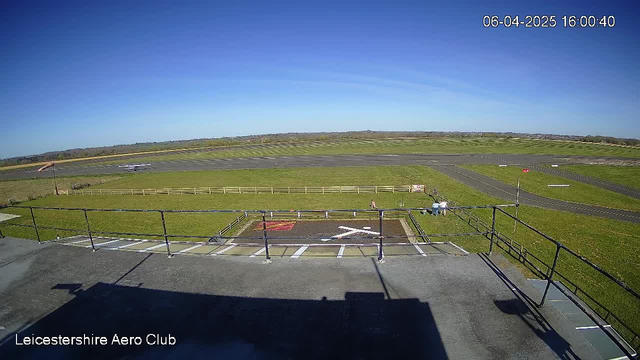 A clear sky with few clouds is visible in the image, showing a grassy area at an airfield. In the foreground, there is a helipad marked with a white cross on a dark surface. A fence surrounds the helipad. In the background, an open runway stretches across the landscape, bordered by green grass and trees in the distance. Two people can be seen near the helipad, and a small plane is positioned further down the runway. The time and date displayed at the top indicate it is 4:00 PM on April 6, 2025.