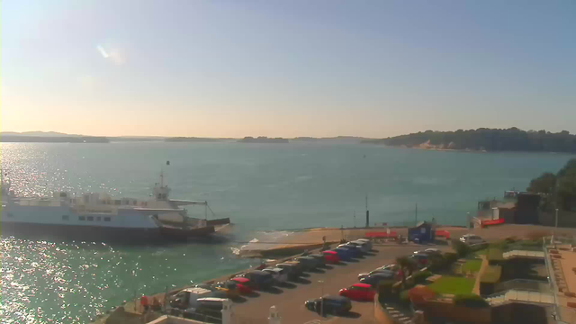 A scenic view of a harbor on a sunny day. In the foreground, there is a parking area filled with cars of various colors. A white ferry boat is docked at the edge of the water, with gentle waves reflecting sunlight. In the background, there's a shoreline lined with trees and distant hills under a clear blue sky. The overall atmosphere is tranquil and inviting.