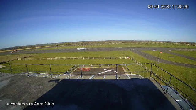 A clear blue sky is visible above a grassy airfield with several runways in the background. In the foreground, a fence outlines a landing area marked with a large white "X." An airplane is positioned on one of the runways in the distance, preparing for takeoff or landing. The setting is serene, with no visible clouds, as the sun casts a bright light over the landscape.