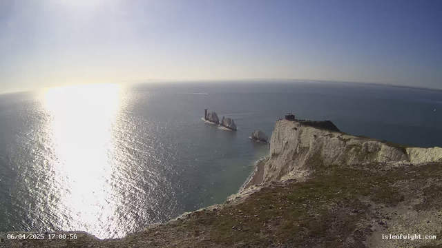 A panoramic view of the ocean under a bright, clear sky. The sun reflects on the water's surface, creating a shimmering effect. In the foreground, there is a cliff with green grass, leading down to the water. An area of white chalk cliffs is visible on the right side of the image. In the distance, several large rock formations rise from the water, and a lighthouse can be seen on one of them. A faint outline of land is visible on the horizon.