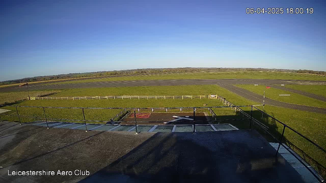 A clear sky with bright sunlight illuminates an expansive landscape. In the foreground, there is a flat rooftop with a railing, casting a shadow. Below, a grassy area leads to an airstrip, which is visible in the mid-ground. The airstrip is bordered by posts and a white fence on the left. To the right, a windsock indicates wind direction. The scene is tranquil, set in an aerodrome environment with no visible aircraft. The timestamp at the top indicates the image was taken on April 6, 2025, at 18:00:19.