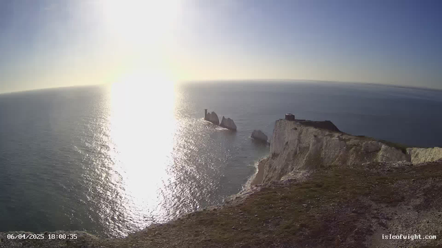 A panoramic view of a coastline during sunset, featuring a bright sun reflecting on calm blue waters. In the foreground, a rocky cliff descends into the ocean, with a small structure atop the cliff. Several large rocks are visible offshore, partially submerged in the water. The sky is clear with a gradient of colors transitioning from a soft blue to golden hues near the horizon.
