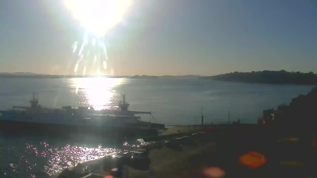 A bright sun is shining in the sky, casting a warm glow over a calm body of water. The sunlight reflects off the water's surface, creating sparkles. In the foreground, a large boat is docked at a pier, with its bow pointing toward the water. Several parked cars are visible on the shore, and a distant shoreline with trees is seen on the horizon. The scene conveys a peaceful, sunny day by the water.